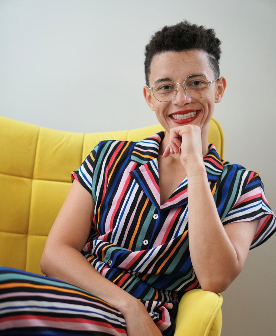 Profile image of the founder, Emma Gardner, facing the camera and smiling. She’s wearing a multi-colored jumpsuit, sitting in a yellow arm chair. She has a short brown afro, wears gold-rimmed glasses, and bright red lip stick.